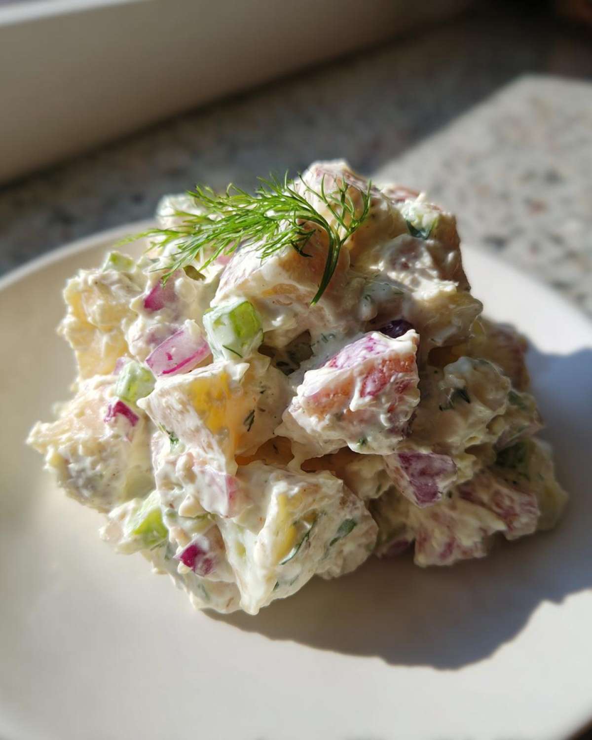 A mound of Creamy Dill Potato Salad featuring chunks of potato, red onion, and celery coated in a white dressing, topped with fresh dill.