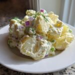 A mound of Creamy Dill Potato Salad featuring chunks of potato, red onion, and peas coated in a rich dressing, garnished with fresh dill.
