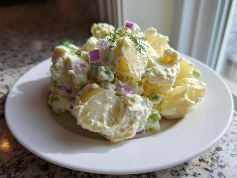 A mound of Creamy Dill Potato Salad featuring chunks of potato, red onion, and peas coated in a rich dressing, garnished with fresh dill.