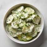 Overhead view of a white bowl filled with slices of Creamy German Cucumber Salad, topped with fresh dill and pepper.