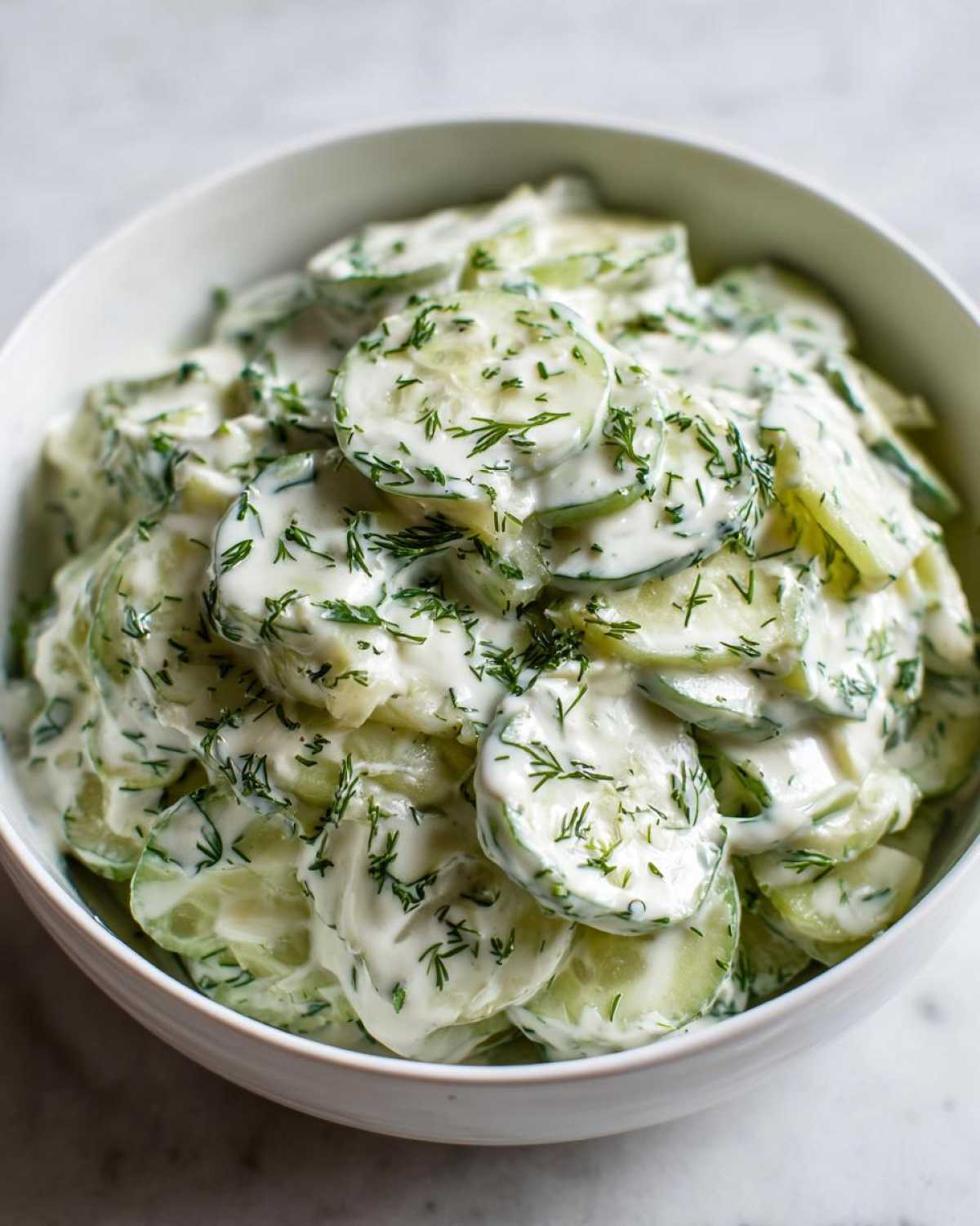 A close-up shot of a white bowl filled with slices of Creamy German Cucumber Salad, generously coated in a white dressing and fresh dill.