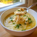 A spoonful of shredded chicken and orzo is lifted from a bowl of Creamy Lemon Chicken Orzo Soup, garnished with parsley.
