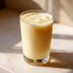 A tall glass filled with a creamy, pale yellow Pineapple Coconut Smoothie, sitting on a marble surface in bright sunlight.