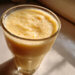 Close-up of a frothy, creamy Pineapple Coconut Smoothie in a tall glass, catching bright sunlight.