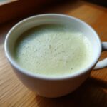 A close-up of a white mug filled with a light green, foamy beverage, likely Creamy Potato Leek Soup, on a wooden surface.