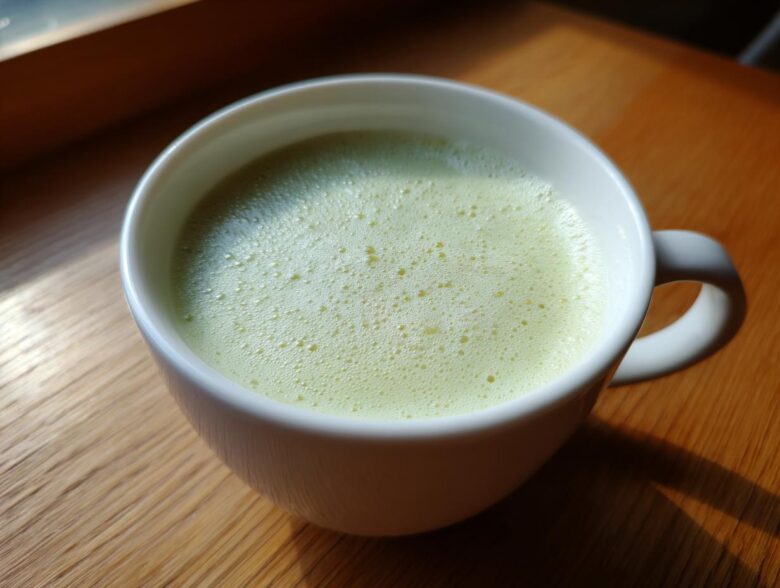 A close-up of a white mug filled with a light green, foamy beverage, likely Creamy Potato Leek Soup, on a wooden surface.
