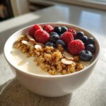 A close-up of a Creamy Vanilla Protein Smoothie Bowl topped with fresh raspberries, blueberries, granola, and sliced almonds.