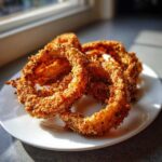 A close-up of several golden brown, crispy Air Fryer Onion Rings piled on a white plate near a window.
