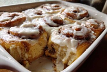 Close-up of warm Gluten-Free Cinnamon Rolls baked together in a white dish, generously covered in white cream cheese icing.
