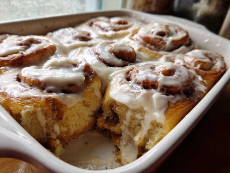 Close-up of warm Gluten-Free Cinnamon Rolls baked together in a white dish, generously covered in white cream cheese icing.