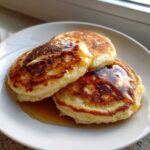 Three fluffy Gluten-Free Buttermilk Pancakes stacked on a white plate, drizzled generously with golden syrup.