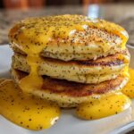 A stack of fluffy Vegan Lemon Poppy Seed Pancakes drizzled with bright yellow lemon sauce and poppy seeds.