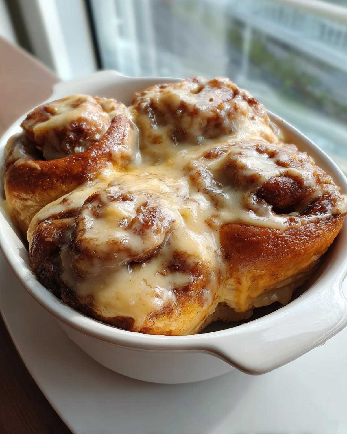 Close-up of four warm Gluten-Free Cinnamon Rolls generously topped with white icing in a white baking dish.