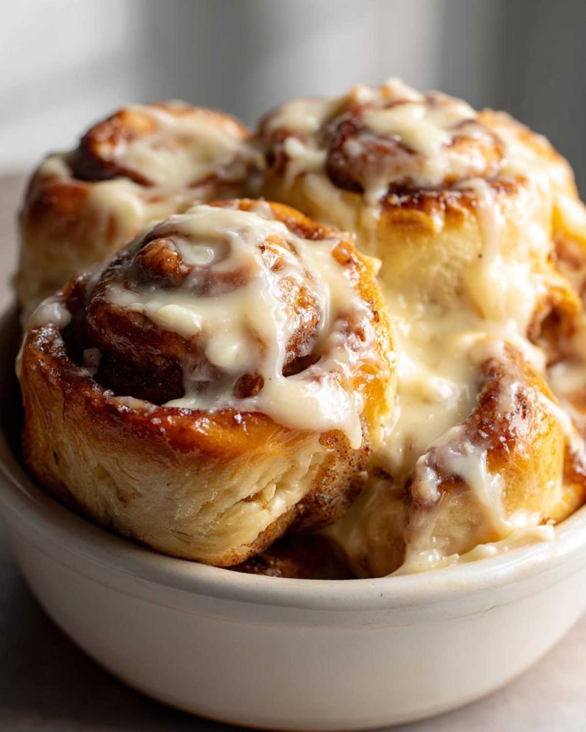 A close-up of several warm Gluten-Free Cinnamon Rolls generously covered in thick white icing, served in a light-colored bowl.