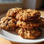 A close-up stack of freshly baked Gluten-Free Oatmeal Raisin Cookies resting on a white plate, illuminated by sunlight.
