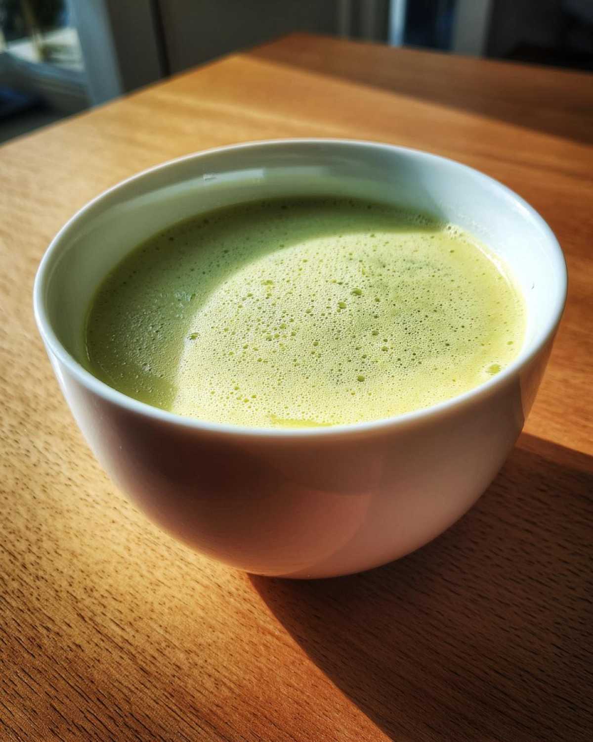Close-up of a bright green, foamy beverage, possibly Creamy Potato Leek Soup, in a white bowl on a wooden table.