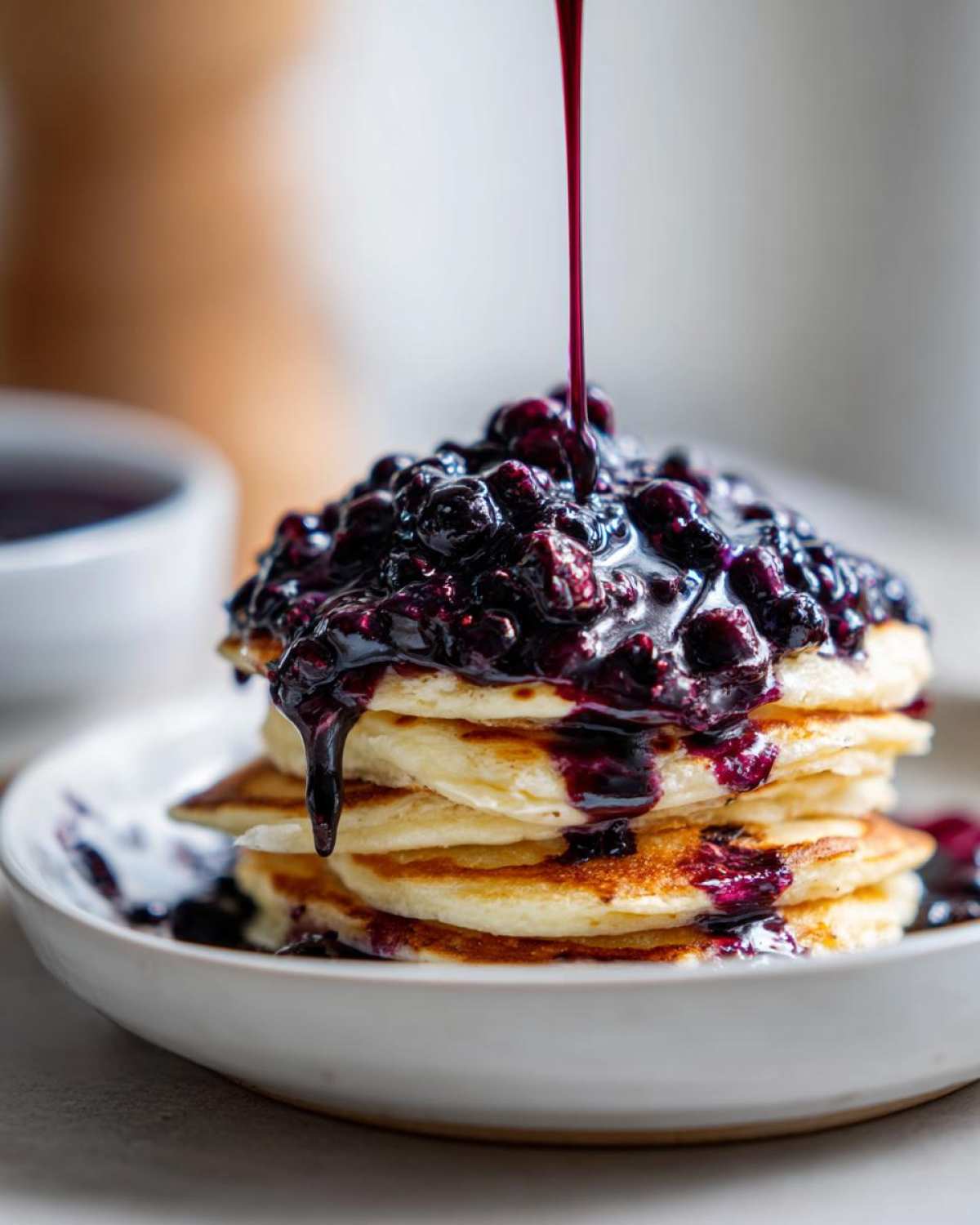 A stream of rich Homemade Blueberry Syrup is being poured over a stack of fluffy pancakes topped with whole blueberries.