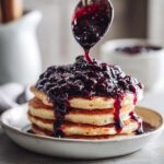 A spoon pours thick Homemade Blueberry Syrup over a stack of fluffy pancakes.