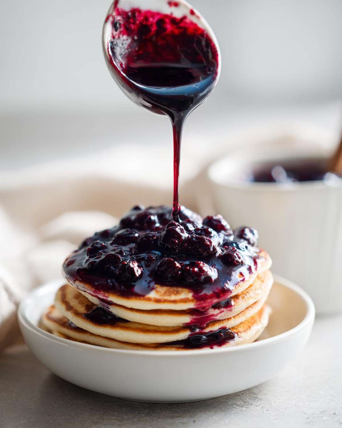 A spoonful of rich Homemade Blueberry Syrup is being poured over a stack of fluffy pancakes.