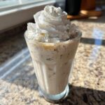 A tall glass of Homemade Vanilla Bean Frappuccino topped generously with whipped cream, sitting on a granite counter.