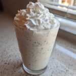 A tall glass of Homemade Vanilla Bean Frappuccino topped high with whipped cream, showing vanilla bean specks.