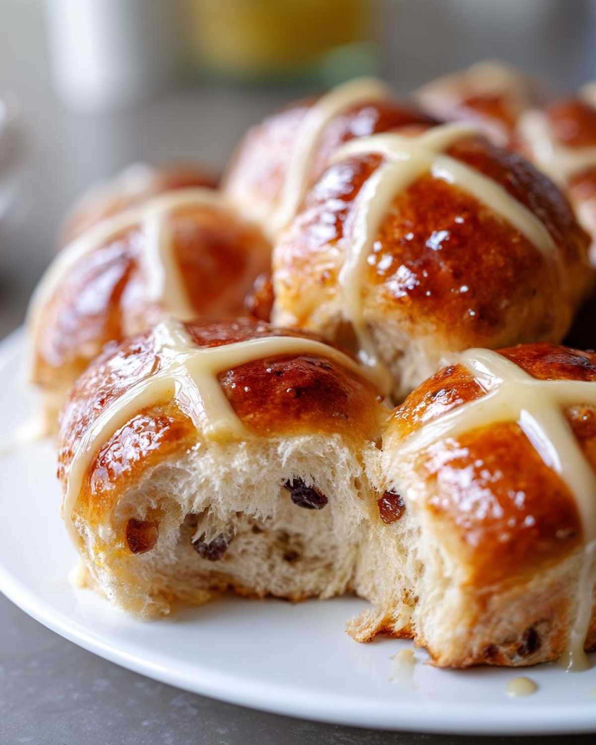 Close-up of soft, glazed Hot Cross Buns with orange glaze, one bun is broken open showing raisins.