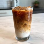 A tall glass of Iced Honey Almond Milk Latte showing layers of white almond milk and dark coffee swirling with ice.