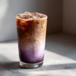A tall glass showing a layered Iced Lavender Latte with purple syrup, milk, and espresso, topped with ice.