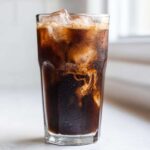 Close-up of a tall glass filled with a dark Iced Mocha, ice cubes, and cream swirling into the coffee.