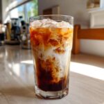 Close-up of a tall glass filled with an Iced Vanilla Chai Latte, showing rich swirls of dark chai concentrate and white milk over ice.