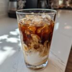 Close-up of an Iced Vanilla Chai Latte showing rich brown tea swirling into white milk over ice cubes.
