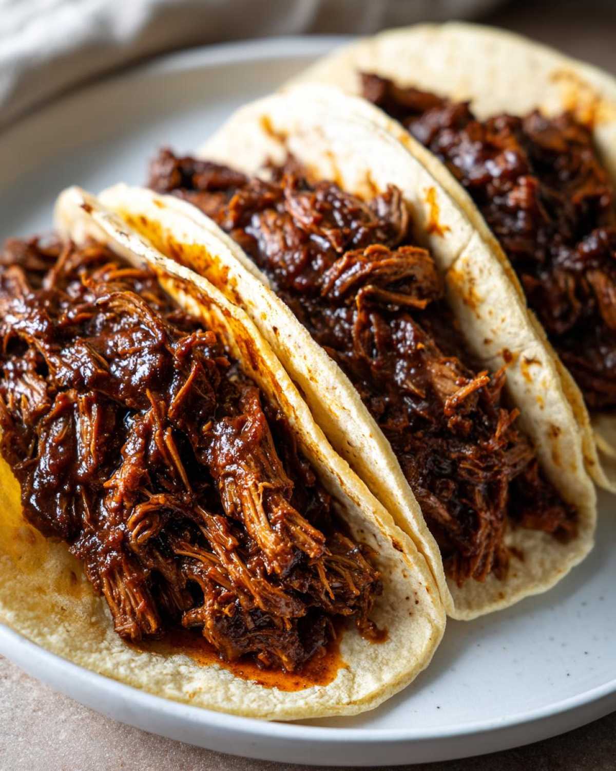 Close-up of three soft corn tortillas filled generously with saucy, shredded Instant Pot Beef Barbacoa.