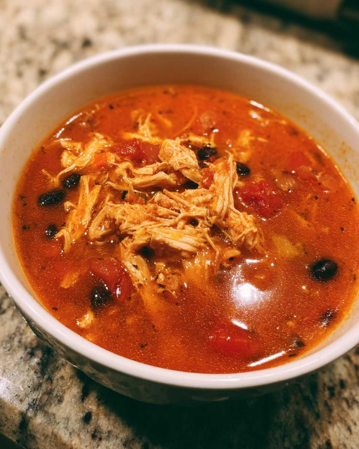 Close-up of a white bowl filled with rich, red Instant Pot Chicken Tortilla Soup topped with shredded chicken and black beans.