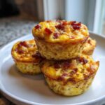A stack of four golden-brown Keto Bacon and Egg Muffins topped with crispy bacon bits on a white plate.