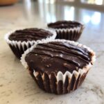 Three delicious Keto Chocolate Peanut Butter Fat Bombs in white paper liners on a light countertop.