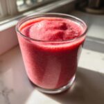 A thick, vibrant pink Keto Strawberry Cheesecake Smoothie served in a clear glass, catching sunlight.
