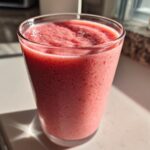 A tall glass filled with a thick, pink Keto Strawberry Cheesecake Smoothie, sitting in bright sunlight.