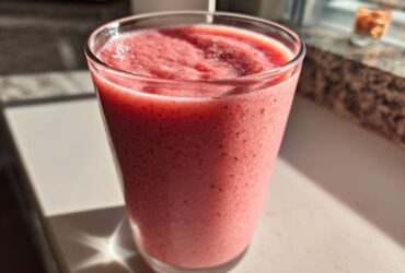 A tall glass filled with a thick, pink Keto Strawberry Cheesecake Smoothie, sitting in bright sunlight.
