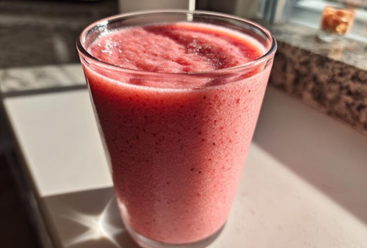 A tall glass filled with a thick, pink Keto Strawberry Cheesecake Smoothie, sitting in bright sunlight.