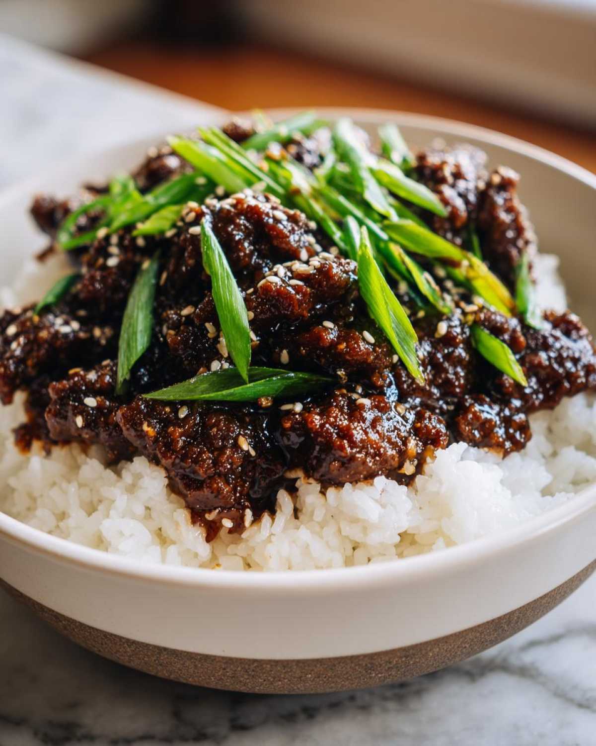 A close-up of a bowl filled with white rice topped generously with saucy Korean Ground Beef Bulgogi, garnished with sesame seeds and sliced green onions.