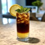 A tall glass showing layers of bright yellow lemonade and dark cold brew coffee over ice, garnished with mint and a lemon slice for a refreshing Cold Brew Lemonade.