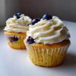 Two Lemon Blueberry Cupcakes topped with swirls of cream cheese frosting and fresh blueberries.