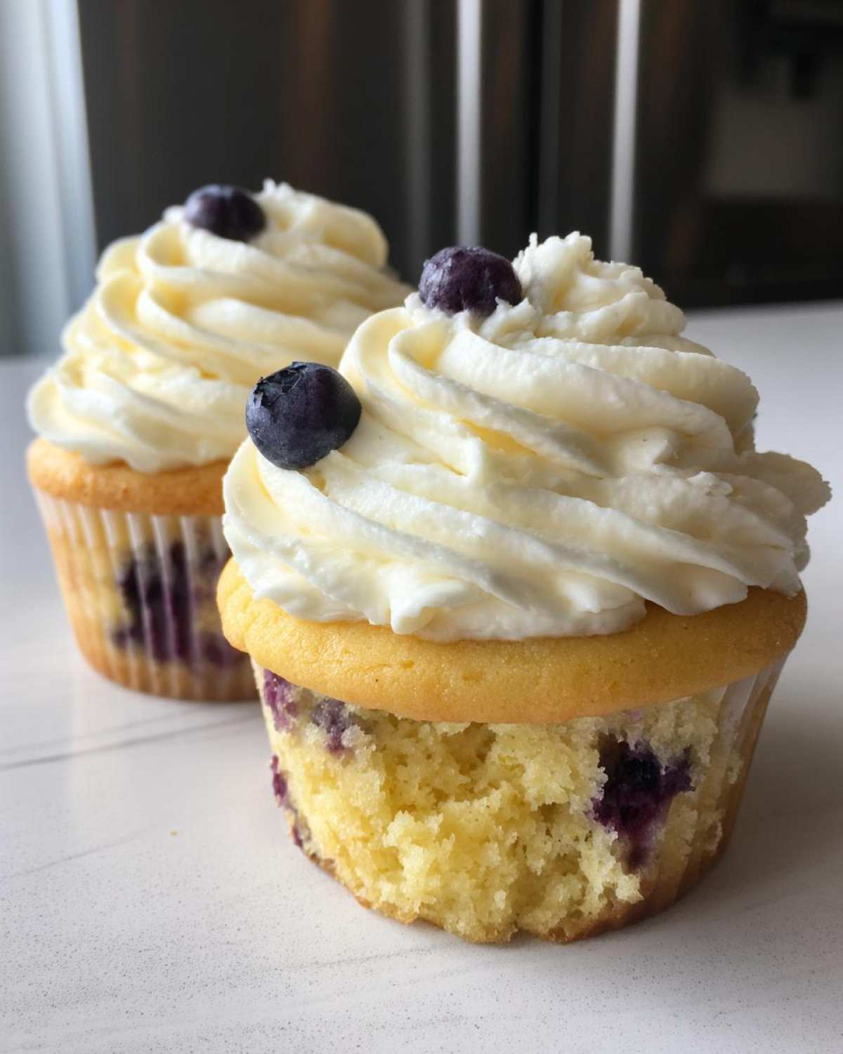 Close-up of two Lemon Blueberry Cupcakes with Cream Cheese Frosting, topped with fresh blueberries.