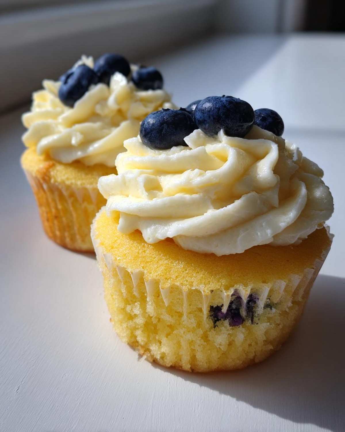 Two Lemon Blueberry Cupcakes topped with swirls of cream cheese frosting and fresh blueberries.