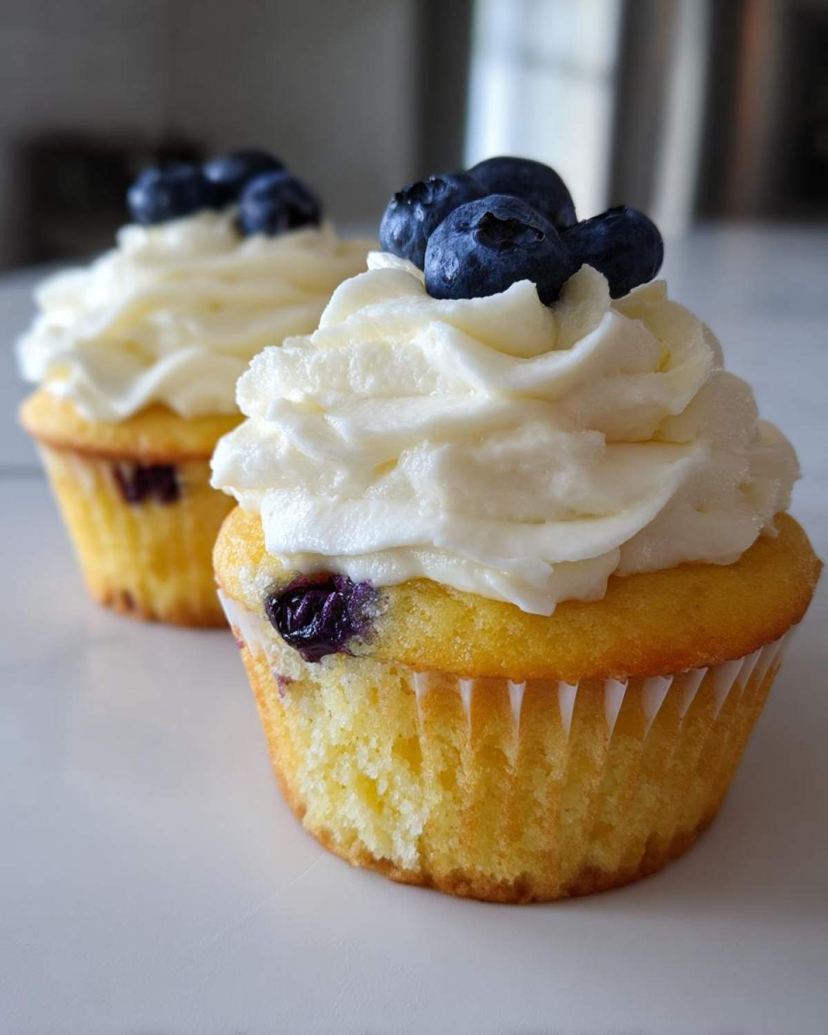 Two Lemon Blueberry Cupcakes with Cream Cheese Frosting, topped with fresh blueberries.