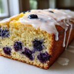 A close-up view of a freshly baked Lemon Blueberry Yogurt Loaf showing moist crumb and dripping lemon glaze.