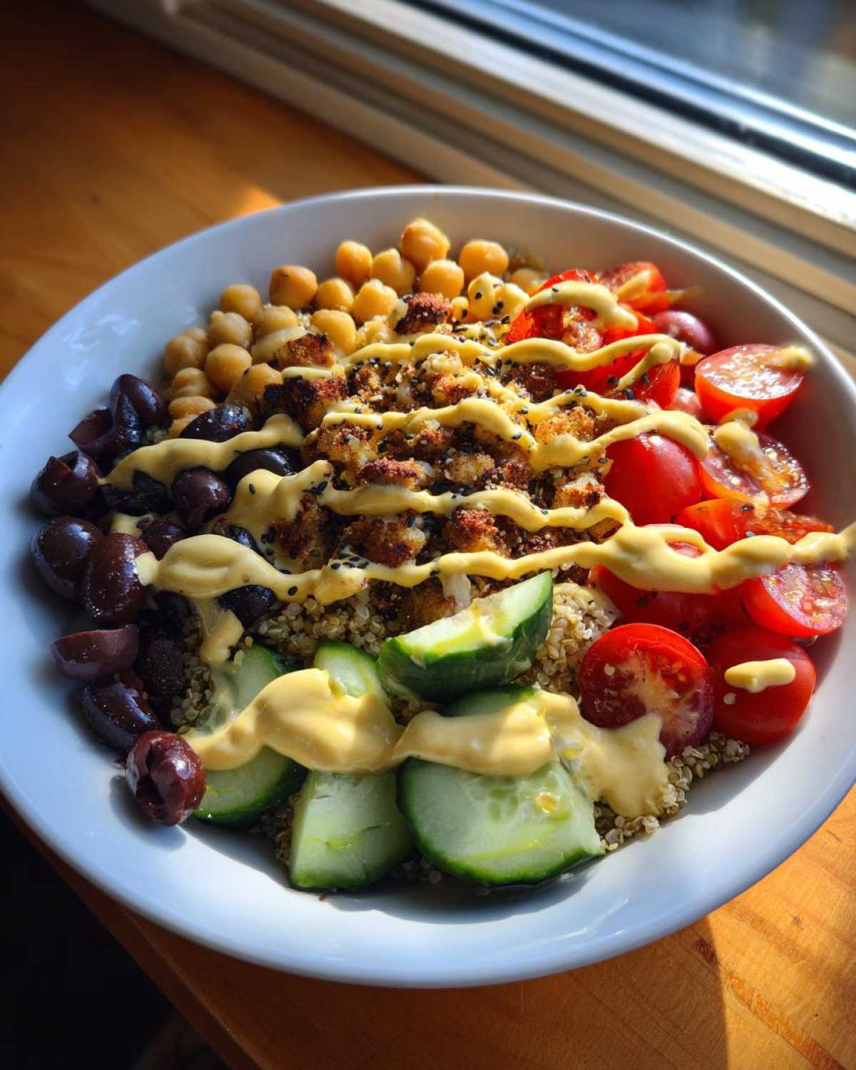 Close-up of a vibrant Mediterranean Quinoa Bowl topped with chickpeas, tomatoes, olives, cucumbers, and drizzled with Lemon Tahini Sauce.
