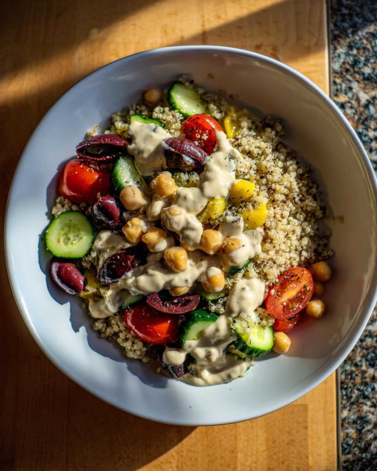 Overhead view of a Mediterranean Quinoa Bowl topped with chickpeas, cucumbers, tomatoes, olives, and lemon tahini sauce.