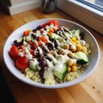 A vibrant Mediterranean Quinoa Bowl topped with cucumbers, tomatoes, olives, chickpeas, and a generous drizzle of lemon tahini sauce.