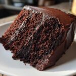 Close-up of a rich, moist slice of Gluten-Free Chocolate Cake covered in thick chocolate frosting on a white plate.